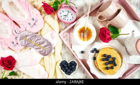 Plateau de petit-déjeuner pancake au lit avec sirop et bleuets fruits mise en page créative plat, vue sur le dessus, avec pantoufles, masque de couchage et chaussettes de lit confortables. Banque D'Images