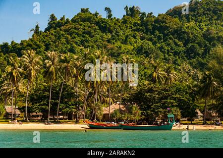 Vue sur l'île de Rabbit, Cambodge de la mer avec des bateaux mordés sur la plage et des touristes étrangers nageant au large de la plage de sable bordée de palmiers. Banque D'Images