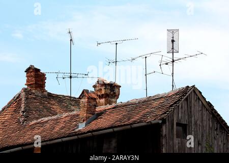 Diverses tailles antennes TV montées sur plusieurs poteaux métalliques sur le toit de maison en bois délabré à côté de cheminées en briques rouges détruites Banque D'Images