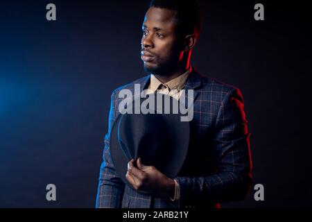 Portrait studio taille haute de jeune homme d'affaires africain peu rasé, confiant et sérieux, portant une chemise, costume à damier debout contre le noirceur Banque D'Images