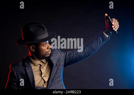 Jeune homme à la mode d'origine africaine, barbu, portant un costume à carreaux, une chemise, un chapeau, un téléphone portable, prenant selfie dans le noir, isolé debout Banque D'Images
