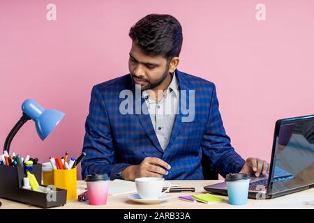 Portrait de jeune entrepreneur indien fatigué pensif assis sur le lieu de travail, tenant le stylo, pensant aux problèmes financiers, porter la combinaison, regarder cependant Banque D'Images