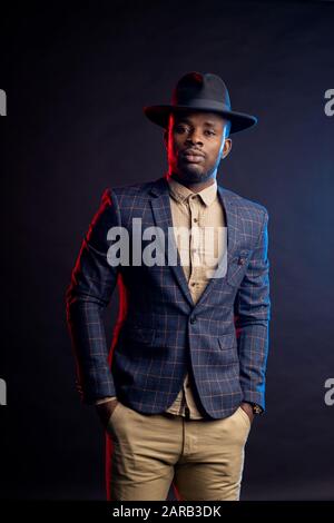 Bel homme posant en studio dans l'obscurité. Portrait d'un gars afro-américain confiant sans rasé portant un costume élégant, chapeau noir debout contre le dos noir Banque D'Images