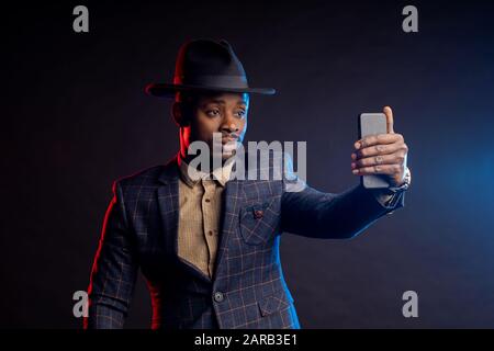Jeune homme à la mode d'origine africaine, barbu, portant un costume à carreaux, une chemise, un chapeau, un téléphone portable, prenant selfie dans le noir, isolé debout Banque D'Images