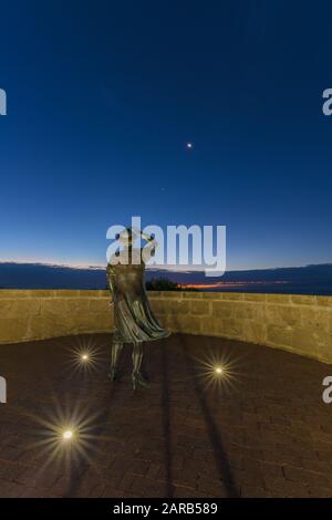 Statue en bronze d'une femme, en soirée, tenant son chapeau dans la brise de la mer surplombant le port de Geraldton sur le site commémoratif du HMAS Sydney 11. Banque D'Images