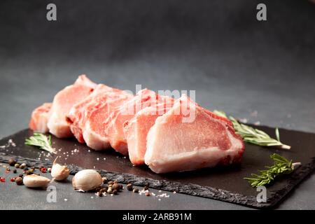 plusieurs steaks de porc crus sur un plateau en pierre noire, épices sur fond noir. Banque D'Images