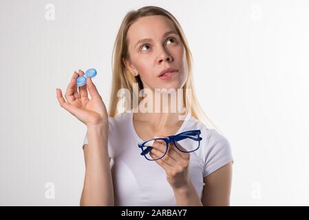 Belle fille blonde choisit entre des lunettes pour la vision ou des lentilles de contact souples sur un fond blanc. Banque D'Images