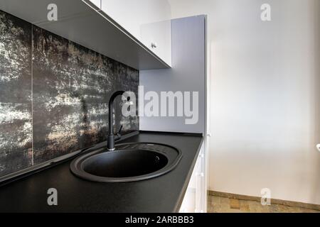 Intérieur d'une cuisine moderne et spacieuse avec mobilier contemporain blanc, carrelage en céramique noire sur le mur et évier en granit sombre avec robinet d'eau. Banque D'Images