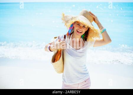 Portrait d'une femme souriante de 40 ans en t-shirt blanc avec sac de paille de plage et grand chapeau sur la plage. Banque D'Images