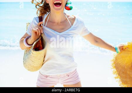 Gros plan sur une heureuse femme moderne dans un t-shirt blanc avec sac de paille de plage et grand chapeau sautant sur la rive de l'océan. Banque D'Images