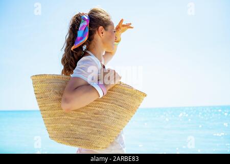 femme en bonne santé en t-shirt blanc avec sac de paille de plage en regardant la distance sur le littoral. Banque D'Images