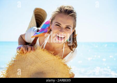 Portrait de heureuse femme moderne d'âge moyen en t-shirt blanc avec sac de paille de plage et grand chapeau sur le bord de mer. Banque D'Images