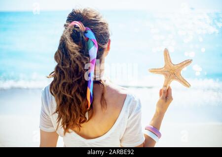 Vue de derrière une femme saine de 40 ans dans un t-shirt blanc avec des étoiles de mer sur la plage. Banque D'Images