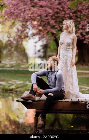 jeune homme avec barbe se trouve sur un pont en bois avec une mariée dans une robe longue de luxe près du lac dans le parc avec des cerisiers en fleurs ou des fleurs de sakura en arrière-plan. Nous Banque D'Images