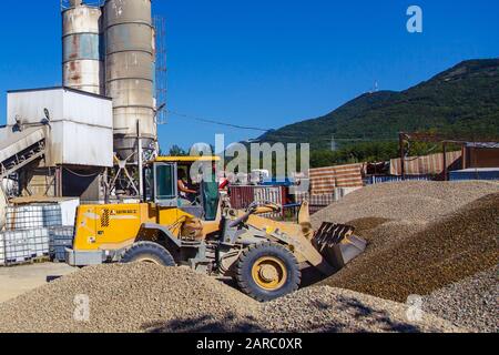 Le chargeur de tracteur charge les décombres lors d'une journée ensoleillée. Piles de décombres. En arrière-plan, l'atelier pour la production de béton. L'équipement du Banque D'Images