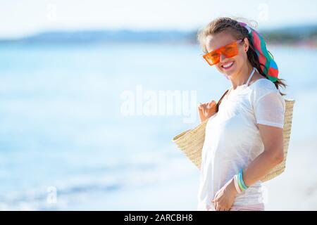 Portrait de heureuse femme moderne de 40 ans en t-shirt blanc et short rose avec sac de paille de plage sur la rive de l'océan. Banque D'Images