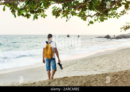 Guy touriste avec un sac à dos jaune marche le long de la plage de sable près de l'océan. Banque D'Images