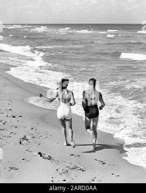 ANNÉES 1930 COUPLE PORTANT DES MAILLOTS DE BAIN JOGGING COURIR ENSEMBLE EN BORDURE DE SURF SUR OCEAN BEACH - B7071 HAR001 HOMME MOYEN-ADULTE MOYENNE-ADULTE FEMME NOIRE ET BLANCHE RACE CAUCASIENNE HAR001 À L'ANCIENNE Banque D'Images