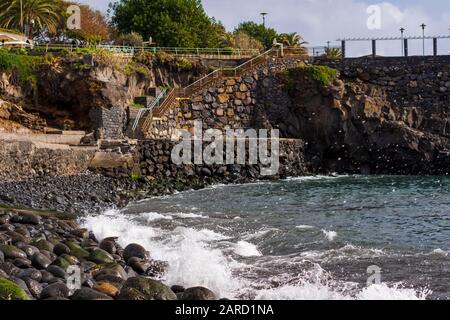 Des vagues éclaboulent vers une plage avec des pierres rondes polies de roche volcanique, et avec un mur et quelques arbres en arrière-plan, photo de Funchal, mdei Banque D'Images