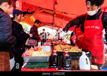 Regardez les gens qui achètent des boîtes en plastique de nouilles dans un étalage de nourriture avec une ligne de cuisiniers servant et préparant la nourriture au festival de la cerisier, au Japon Banque D'Images