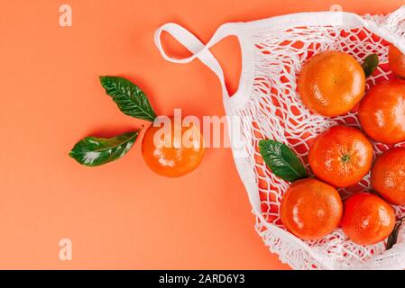 Mandarines fraîches mûres avec feuilles vertes dans un sac en coton blanc sur fond orange. Plat, vue du dessus. Banque D'Images