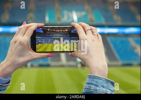 Une fille dans un stade de football (football) avec un smartphone prend une photo du champ vert de l'arène. Technologie moderne lors d'événements sportifs. Banque D'Images