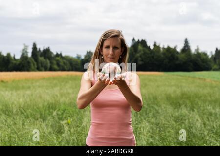 Vue de face d'une jeune femme méditant dans la nature tenant la boule de cristal dans ses mains avec prairie et forêt en arrière-plan. Banque D'Images