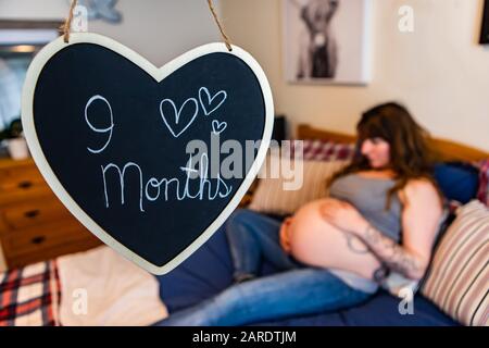 Un foyer sélectif d'un signe de tableau de surveillance cardiaque disant 9 mois. Avec une femme enceinte floue posée dans le lit se reposant les mains sur le bébé bosse en arrière-plan Banque D'Images