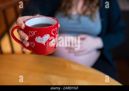 Une femme enceinte est vue profiter d'un café du matin avec une faible profondeur de champ et une bosse floue de bébé en arrière-plan, servi dans la tasse d'amour rouge, copier l'espace à droite Banque D'Images