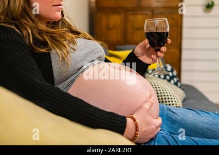 Une femme très enceinte approchant le travail est vue assise sur un canapé tenant tummy gonflé d'une main et un verre de vin rouge dans l'autre, avec espace de copie Banque D'Images