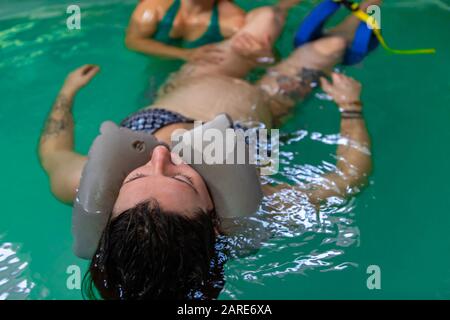 Jeune femme enceinte sur costume bleu de natation allongé dans la piscine avec le cou flotter autour de la tête et la thérapeute féminine aidant à l'exercice des jambes Banque D'Images