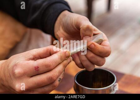 Gros plan sélectif de la mise au point de mains tatouées tordant un papier avec de la marijuana moulu préparant un cannabis sous une bouteille de métal sur une table à damier. Banque D'Images