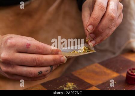 Foyer sélectif gros plan de mains tatouées remplissant un papier roulant avec de la marijuana moulu préparant un joint de cannabis sur une table à damier. Banque D'Images