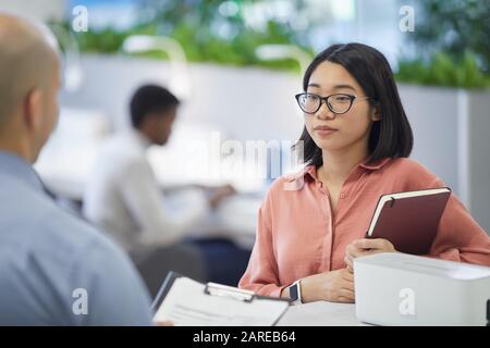 Portrait de la jeune femme d'affaires asiatique parlant au patron ou au Manager tout en se tenant dans l'intérieur moderne de bureau, espace de copie Banque D'Images