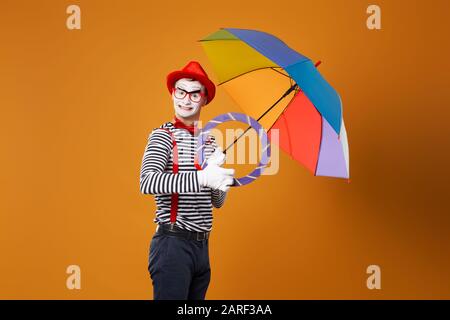 Mime drôle avec parapluie multicolore sur fond orange vide en studio Banque D'Images