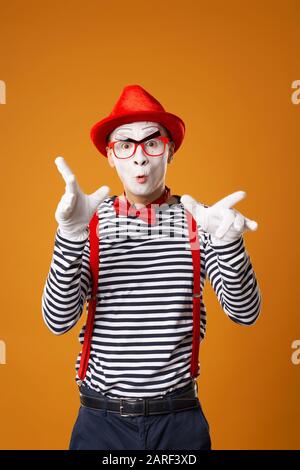 Jeune mime avec des gants blancs et un chapeau rouge regardant l'appareil photo sur fond orange vide Banque D'Images