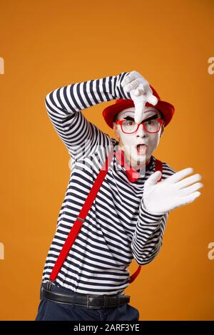 Homme de mime sérieux avec des gants blancs et un chapeau rouge regardant l'appareil photo sur fond orange vide Banque D'Images