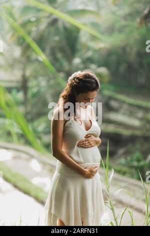 Jeune femme enceinte en robe blanche avec vue sur les rizières en terrasses de Bali dans la lumière du soleil du matin. Harmonie avec la nature. Concept de la grossesse. Banque D'Images