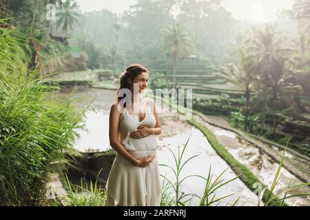 Jeune femme enceinte en robe blanche avec vue sur les rizières en terrasses de Bali dans la lumière du soleil du matin. Harmonie avec la nature. Concept de la grossesse. Banque D'Images