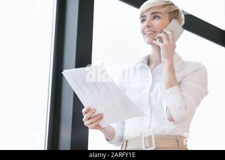 Femme D'Affaires Parlant Sur Cellphone Debout Près De La Fenêtre Dans Le Bureau Moderne Banque D'Images