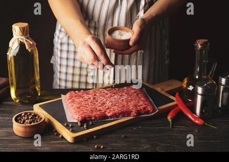 Femme saupoudrez de sel sur de la viande hachée sur une table en bois avec des épices, gros plan Banque D'Images