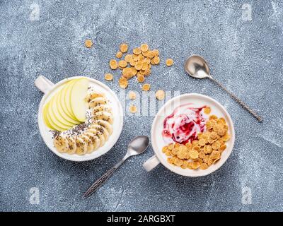 Deux yaourts grecs avec confiture, pomme, muesli, graines de chia et banane dans un bol blanc sur table en pierre de béton bleu gris, vue de dessus Banque D'Images