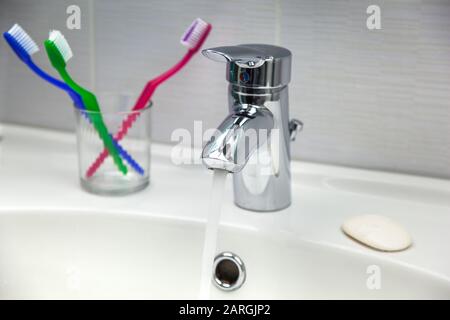 Brosses à dents, savon et robinet de robinet d'eau avec eau courante propre dans un évier de salle de bains blanc. Banque D'Images