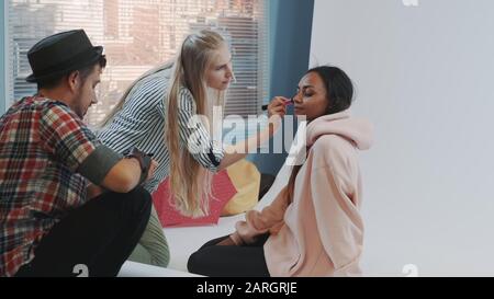 Gros plan de maquillage artiste appliquant maquillage sur beau modèle noir assis sur le sol et posant. Photographe prenant des photos sur un appareil photo professionnel. Banque D'Images