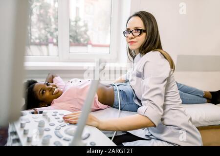 Jeune femme souriante médecin déplaçant la sonde à ultrasons sur le ventre et les reins d'une femme africaine tout en regardant l'écran à l'hôpital Banque D'Images