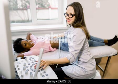Jeune femme souriante médecin déplaçant la sonde à ultrasons sur le ventre et les reins d'une femme africaine tout en regardant l'écran à l'hôpital Banque D'Images