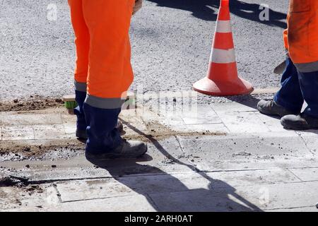 Jambes de deux travailleurs en pantalon uniforme orange, réparation de chaussée le jour ensoleillé Banque D'Images