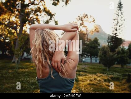 Vue arrière de la jeune femme blonde dans des vêtements de sport qui étirent son bras dans le parc - femme sportive qui s'étend dans le parc Banque D'Images