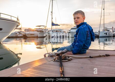 Un garçon dans une veste bleue se trouve sur une jetée avec des yachts au coucher du soleil. Beau coucher de soleil clair dans le port de mer avec des yachts amarrés. Banque D'Images