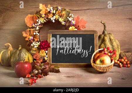 Arrangement d'automne avec couronne et lettrage 'Happy Fallidays' sur un tableau de craie. Panier de pommes et de baies, citrouilles décoratives, feuilles d'automne et Banque D'Images